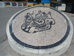 Foto do símbolo da escola que está reproduzido com através da calçada portuguesa no dos espaços da escola.